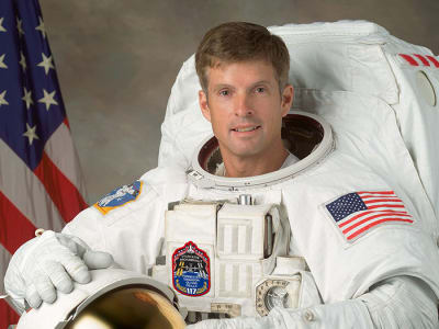Headshot of astronaut Steve Swanson in full space suit holding his helmet in front of the U.S. flag