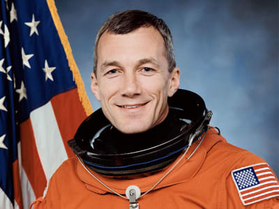 Headshot of astronaut Terry Wilcott in orange space suit minus helmet in fron the of the U.S. flag