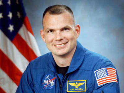 Fotografía del astronauta Tony Antonelli con traje de vuelo azul delante del fla de EE.UU.
