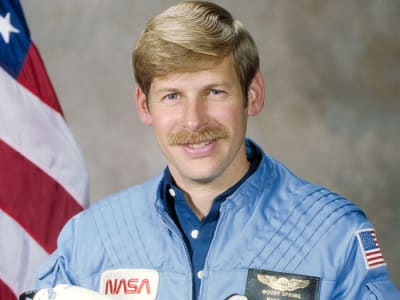 Headshot of astronaut Woody Spring in blue flight suit holding a model of the space shuttle in front of the U.S. flag