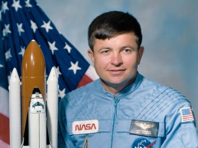 Headshot of astronaut Ken Cameron in blue flight suit in front of the U.S. flag and next to a model of the space shuttl