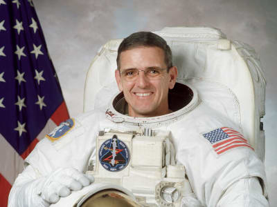 Headshot of astronaut Bill McArthur in space suit holding his helmet in front of U.S. flag
