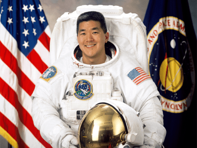 Astronaut Daniel Tani in white flight suit holding his helmet in front of the American flag and NASA flag