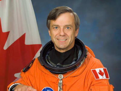 Astronaut Robert “Bob” Thirsk wearing an orange flight suit and standing in front of a Canadian flag