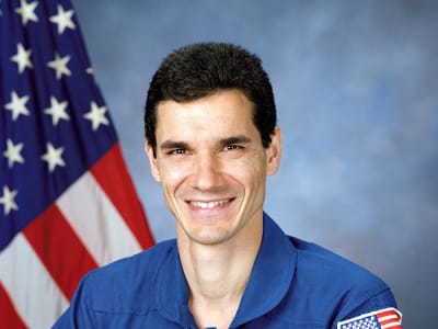 Headshot photo of NASA astronaut Gregory Linteris wearing a blue flight suit in front of the American flag