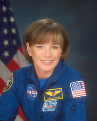 Fotografía de la astronauta Anna Fischer con un mono azul de la NASA y la bandera de EE.UU. de fondo.