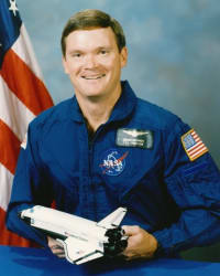 Astronaut Bruce Melnick in blue flight suit standing with arms crossed in front of space shuttle Atlantis