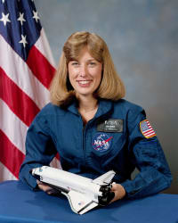 Kopfbild des Astronauten Jan Davis im blauen Fluganzug, der ein Modell eines Spaceshuttles vor der US-Flagge hält.