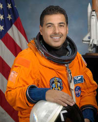 Kopfbild des Astronauten José Hernandez im orangefarbenen Raumanzug, der seinen Helm hält und vor der US-Flagge und einem Modell des Space Shuttle steht
