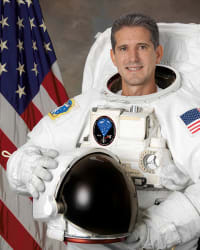 Kopfbild des Astronauten Mike Good in vollem Raumanzug mit Helm vor der US-Flagge.