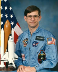 Foto des NASA-Astronauten Robert Cenker neben der Flagge der Vereinigten Staaten und einem Modell des Spaceshuttles