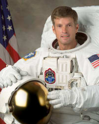 Headshot of astronaut Steve Swanson in full space suit holding his helmet in front of the U.S. flag