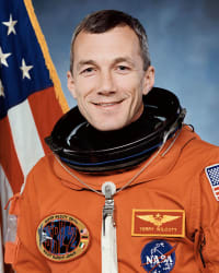Headshot of astronaut Terry Wilcott in orange space suit minus helmet in fron the of the U.S. flag