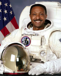 Kopfbild des Astronauten Winston Scott in vollem Raumanzug, der seinen Helm hält und vor der US-Flagge steht.