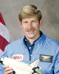 Kopfbild des Astronauten Woody Spring im blauen Fluganzug, der ein Modell der Raumfähre vor der US-Flagge hält.