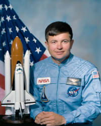 Headshot of astronaut Ken Cameron in blue flight suit in front of the U.S. flag and next to a model of the space shuttl