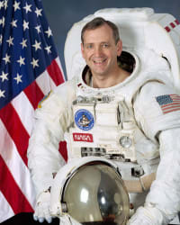 Fotografía oficial de la NASA del astronauta Tom Akers, vestido con un traje espacial y con la bandera estadounidense al fondo