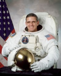 Istantanea dell'astronauta Bill McArthur in tuta spaziale con il casco in mano davanti alla bandiera degli Stati Uniti.