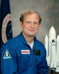 Tête de l'astronaute Norman Thagard en combinaison de vol bleue devant le drapeau de la NASA et la maquette de la navette spatiale.