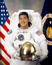 Astronaut Daniel Tani im weißen Fluganzug hält seinen Helm vor der amerikanischen Flagge und der NASA-Flagge