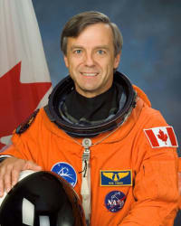Astronaut Robert “Bob” Thirsk wearing an orange flight suit and standing in front of a Canadian flag