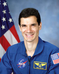 Foto frontale dell'astronauta della NASA Gregory Linteris che indossa una tuta di volo blu davanti alla bandiera americana