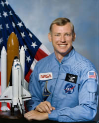 Fotografía del astronauta Mark Brown con traje de vuelo azul delante de la bandera de EE.UU. y junto a una maqueta del transbordador espacial.