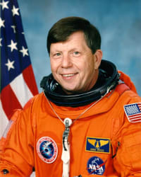 Tête de l'astronaute Roger Crouch en combinaison spatiale orange sans casque devant un drapeau américain.