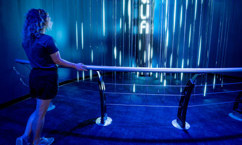 A woman with blonde hair leans on a circular railing as she looks inward at lights pulses on vertical beams.