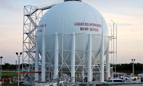 A large, round, white liquid hydrogen tank with vertical white support structures. The words "Liquefied Hydrogen MAWP: 90 PSID" are written across it in red.