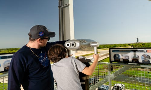 A brown-haired boy of 12 years wearing a grey t-shirt and his father, wearing a long-sleeved navy shirt with NASA logo and a grey ballcap with NASA logo are standing at the edge of the third floor balcony of the Gantry, looking outward at Launch Pad 39A The boy is holding onto a metal pair of viewing binoculars, while his father points into the distance. The boy is leaning towards him, as if to see what he is pointing at. In the distance on the launch pad is the SpaceX launch tower.