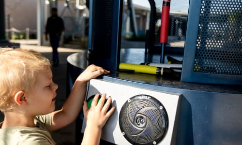 A blonde boy around six years old hovers his hand over a green button on a cylindrical metal console. Atop the console, a red Styrofoam rocket stands upright on a pole.