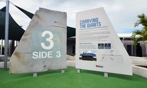 A large, grey metal piece of the Mobile Launch Platform, about twelve feet tall by twelve feet wide with a large number three and the words "Side 3" printed on it, stands next to a mirrored piece of the same shape. The mirrored piece has information about the artifact on it, relaying its history and importance.