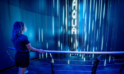 A woman with blonde hair leans on a circular railing as she looks inward at lights pulses on vertical beams.