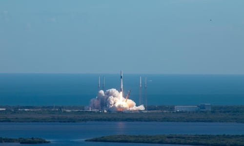 A distant shot of a SpaceX Falcon 9 rocket launching from Cape Canaveral. Smoke plumes below the rocket and the launch tower falls towards the ground. The Atlantic Ocean is in the background.