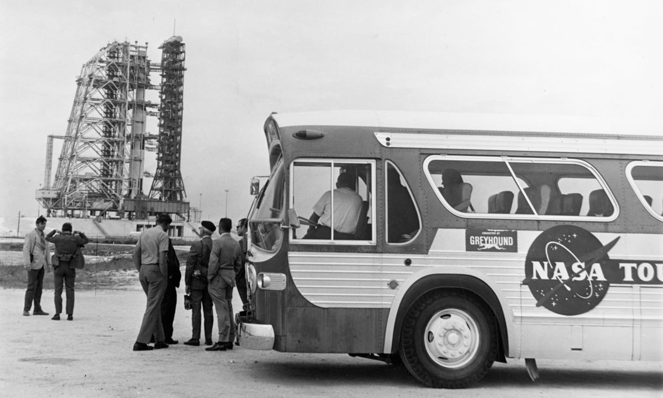 This photograph belongs to the Florida Memory State Library and Archives collection, documenting a public tour stop at the Kennedy Space Center in July 1967. his particular photo highlights a GMC New Look Suburban bus operated by Greyhound, featuring a distinctive "Conducted by Greyhound" placard right next to the circular NASA Tours decal. he bus is parked at a visitor observation point near Launch Complex 34 or Launch Complex 39, giving tourists an up-close look at the massive service gantry and mobile launcher tower structures visible in the background.