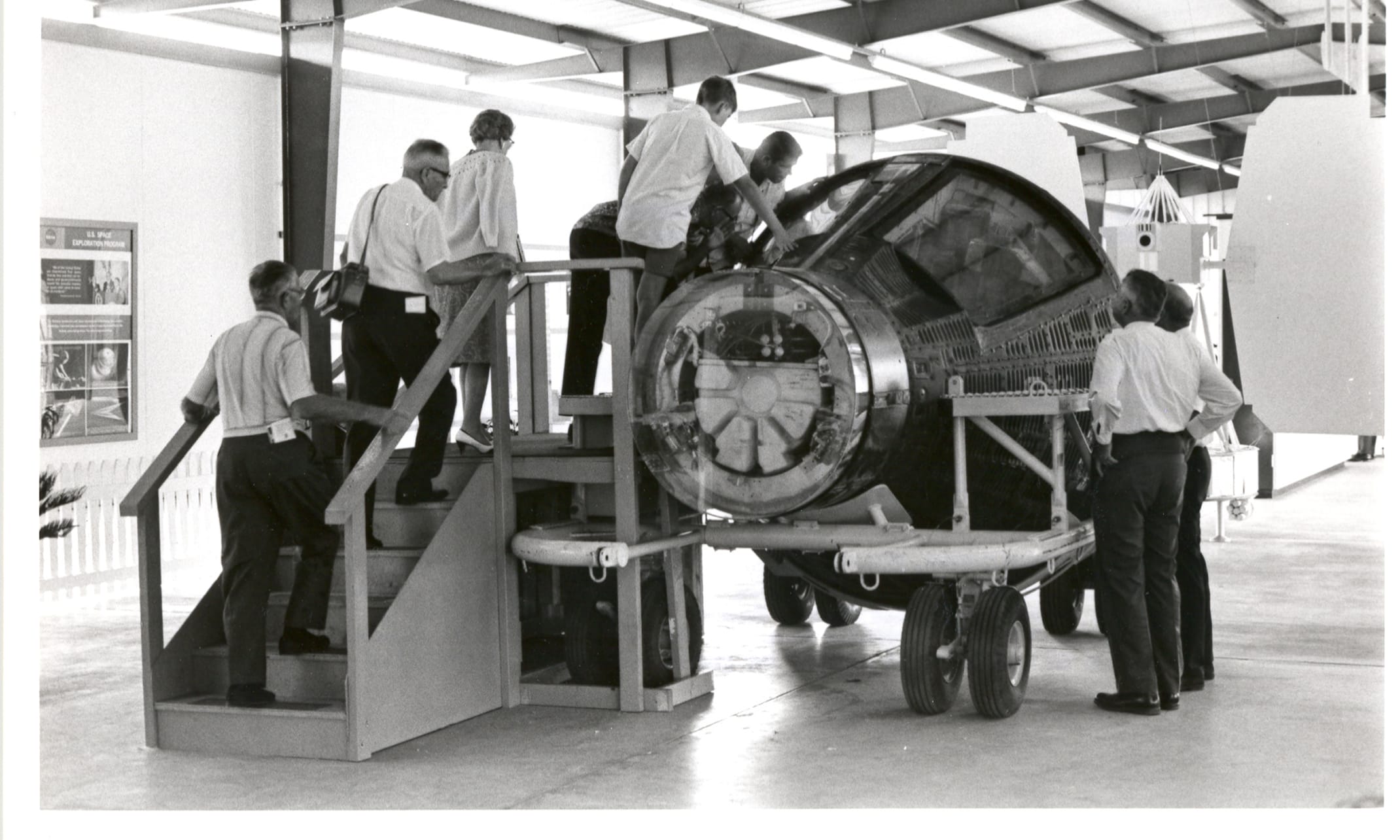 This photograph is from the official NASA Kennedy Space Center digital archive, taken in late 1967. It documents the public exhibition of space flight hardware inside the newly opened Visitor Information Center (VIC). Visitors are climbing a wooden staircase to look inside a flown Project Gemini spacecraft capsule. The capsule is mounted on a mobile transport cradle, with its rear adapter sections detached to let the public view the complex wiring, interior instrumentation, and astronaut seating arrangement. This scene took place inside the display bays of the permanent Visitor Information Center, which officially opened its doors to the public on August 1, 1967. This facility marked a major upgrade from the single roadside information trailer used in 1966.