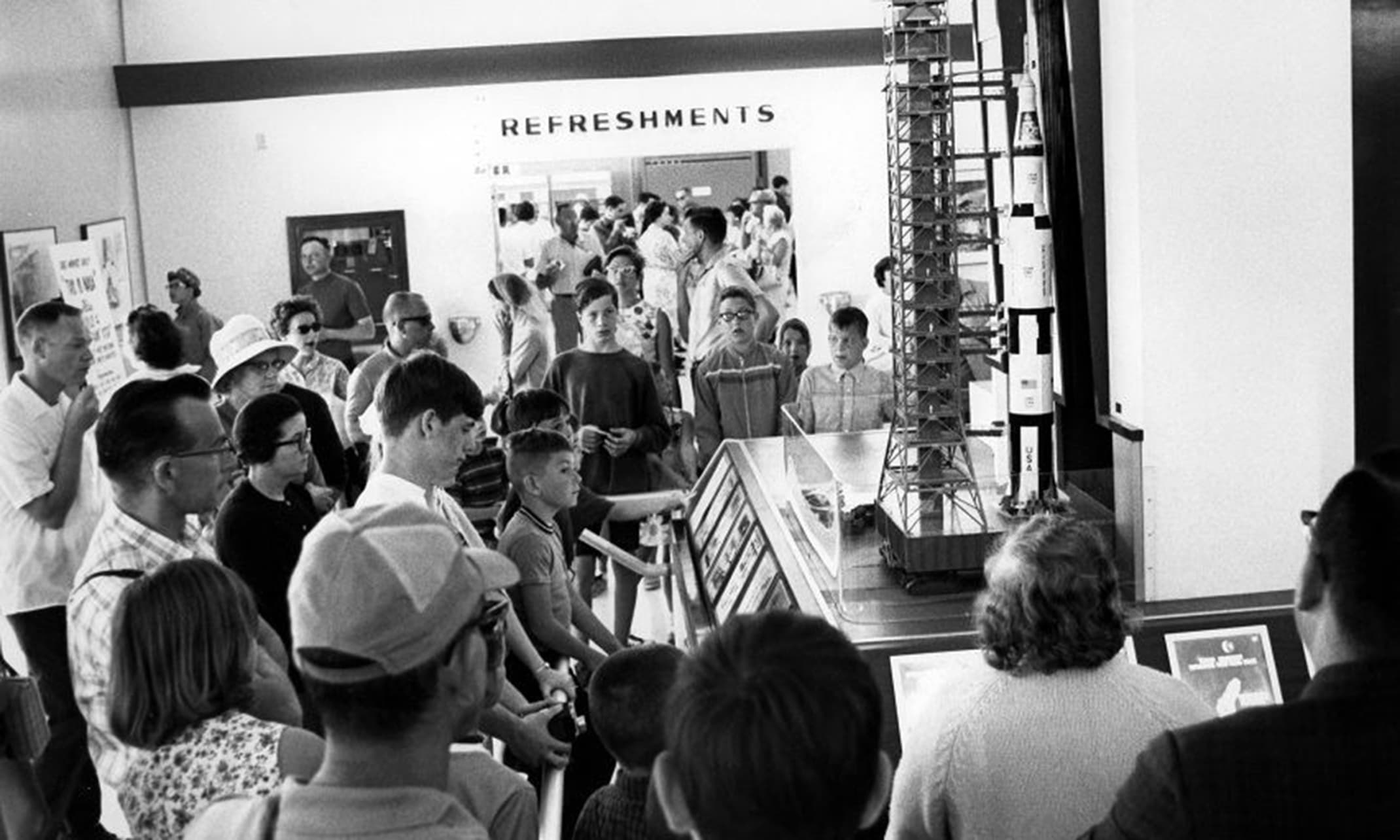 This photograph is from the official NASA Kennedy Space Center digital archive, captured in late 1967 inside the newly opened Visitor Information Center (VIC). Visitors are gathered around an interactive, highly detailed scale model of a Saturn V rocket standing alongside its Mobile Launcher tower and umbilical service arm structure. This scene took place inside the display hall of the permanent Visitor Information Center, which opened on August 1, 1967. In the background, the prominent sign for the facility's "REFRESHMENTS" hub and snack bar is visible, bustling with early spaceport tourists. he photo captures the public excitement of the late 1960s Apollo era, giving regular citizens an up-close look at the massive engineering scale of the hardware planned to take astronauts to the moon.