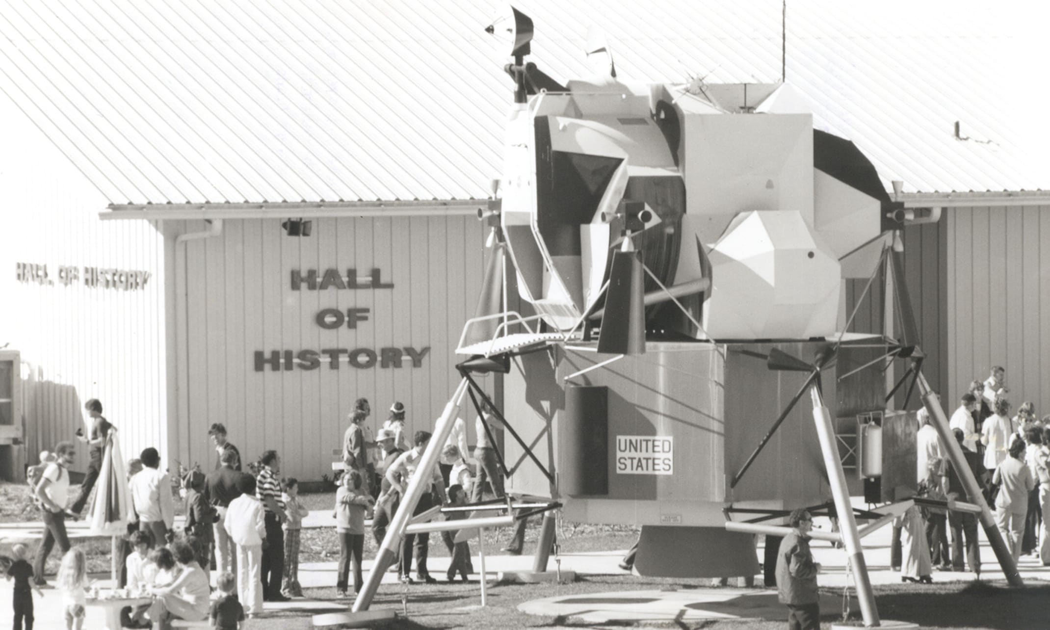 This photograph comes from the official NASA Kennedy Space Center digital archive, capturing a popular outdoor display from the mid-to-late 1970s. The centerpiece of this courtyard display is a full-scale engineering mockup of an Apollo Lunar Module (LM). It features a detailed descent stage, complete with landing pads, a surface ladder, and the upper crew ascent stage cabin. The photo was taken around 1976, catching a heavy crowd of tourists exploring the grounds during the peak of America's Bicentennial celebration. This particular display area eventually evolved into the world-famous Rocket Garden seen by visitors today.