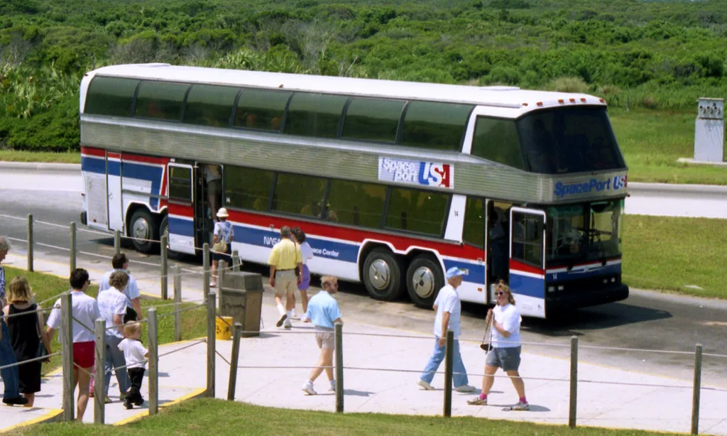 In July 1985, NASA's visitor complex debuted a fleet of custom-built, high-capacity double-decker coaches. This photograph comes from the official Kennedy Space Center archive and Alamy's historical stock photo library, capturing visitors boarding a tour coach in the late 1980s or early 1990s. The bus pictured is an early Neoplan Super Skyliner (N128/4). Custom-manufactured by NEOPLAN USA, these massive four-axle, 47-foot-long transport vehicles featured a distinct 8x2 drivetrain configuration and held up to 106 passengers simultaneously across two levels. The coach sports the red, white, and blue stripe scheme of "Spaceport USA," which was the official marketing name for the Kennedy Space Center Visitor Complex from the launch of the Space Shuttle program until it was rebranded in the late 1990s.