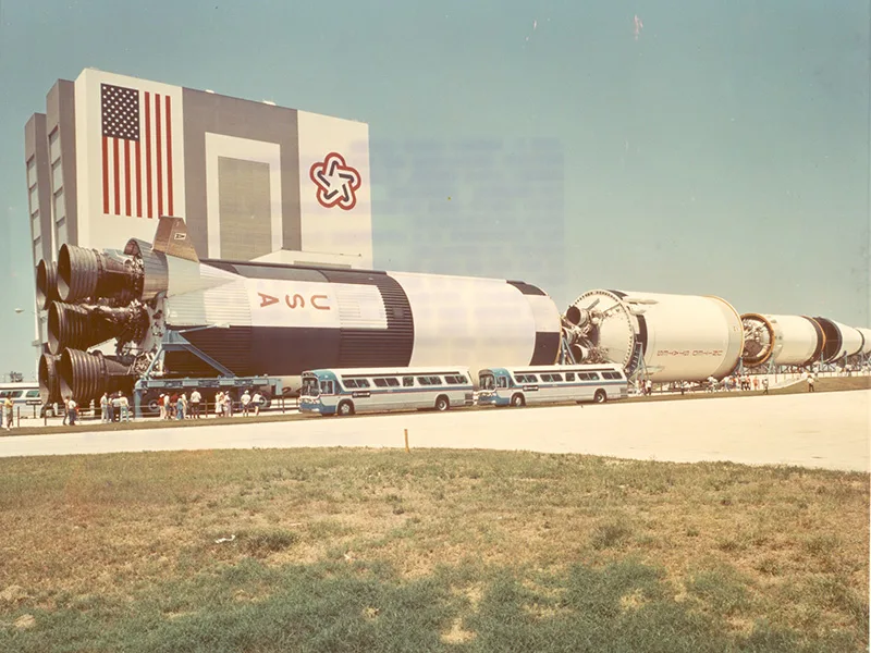 This photograph comes from a postcard documenting the outdoor display of a Saturn V rocket in front of the Vehicle Assembly Building (VAB) at Kennedy Space Center. The distinctive red, white, and blue U.S. Bicentennial Star logo was painted on the side of the VAB to celebrate America's 200th birthday. (1976)