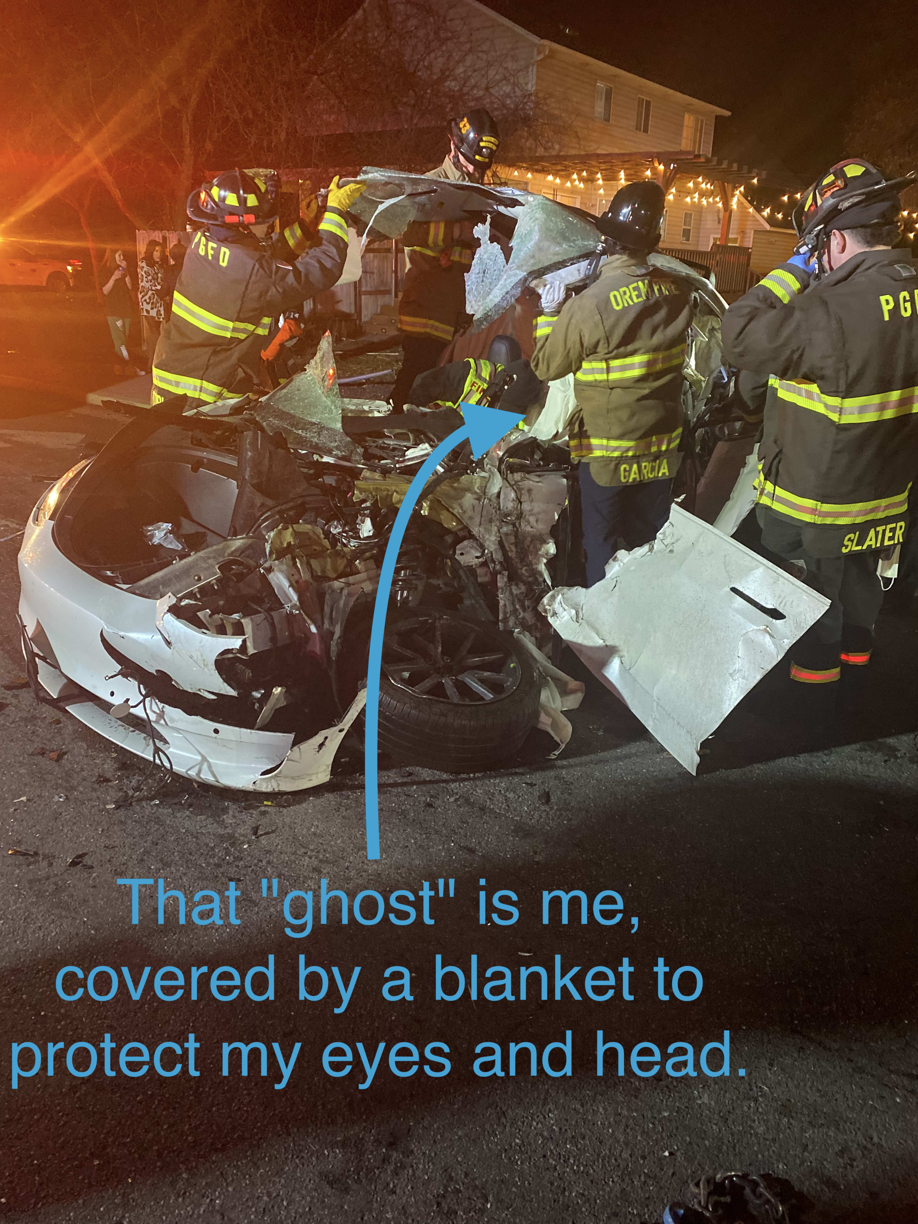 A destroyed Tesla model 3 with firefighters around helping to extract Kent who is covered in a white sheet. An arrow is pointed at Kent with the words: That "ghost" is me, covered by a blanket to protect my eyes and head.