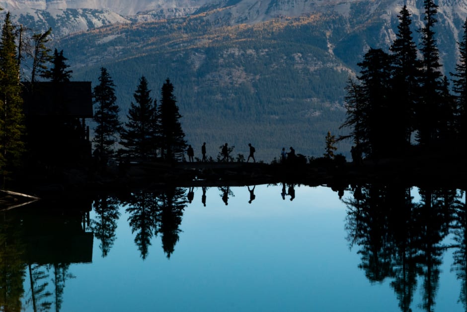 A silhouette of hikers next to Lake Agnes tea house with the reflection of the lake in front of them.