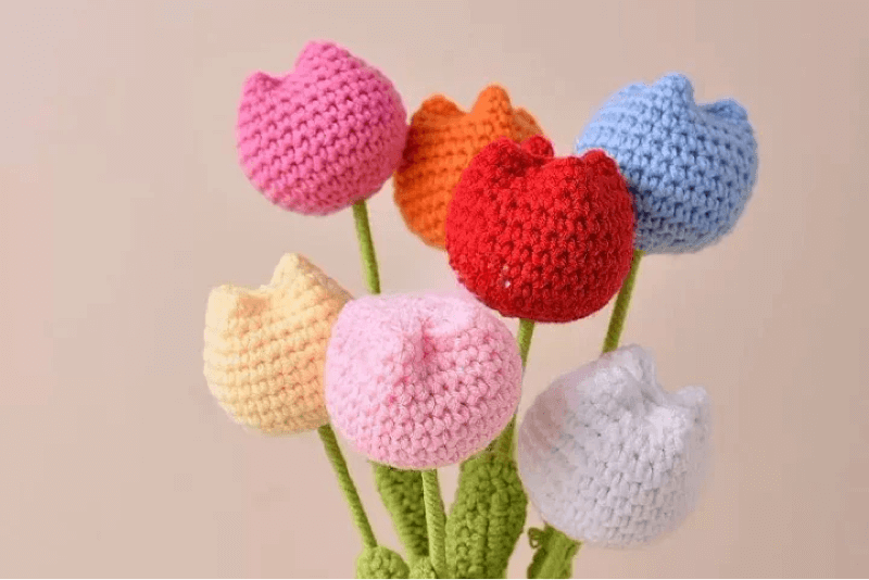 Handmade crochet tulip bouquet featuring colorful knitted flowers in red, pink, yellow, orange, blue, and white, with green stems, set against a soft pastel background.