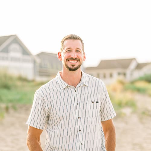 Peter Kirk smiling while on white sandy beach in a striped button up shirt.