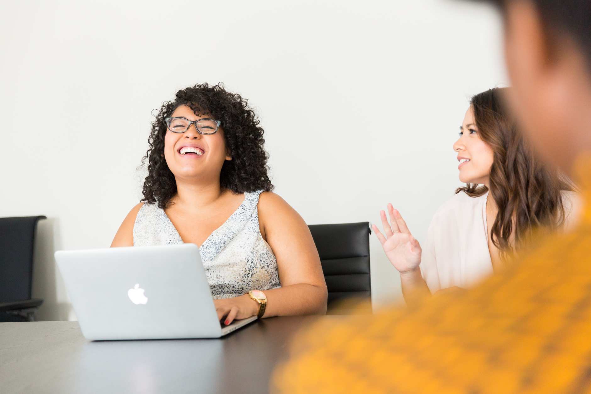 an image showing a woman laughing while working on a laptop and talking to another woman 