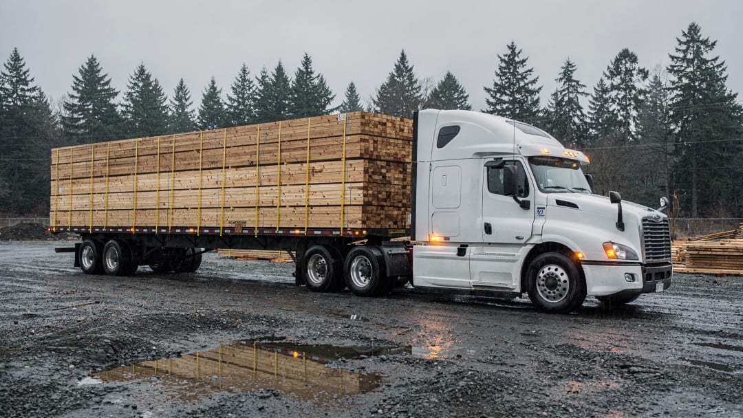 A white tractor with a flatbed of strapped lumber at a gravel job site on an overcast day. The kind of equipment we run.