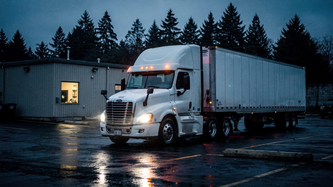 A white tractor and dry van at a small truck yard at blue hour, wet asphalt and one office window lit. The kind of equipment we run.