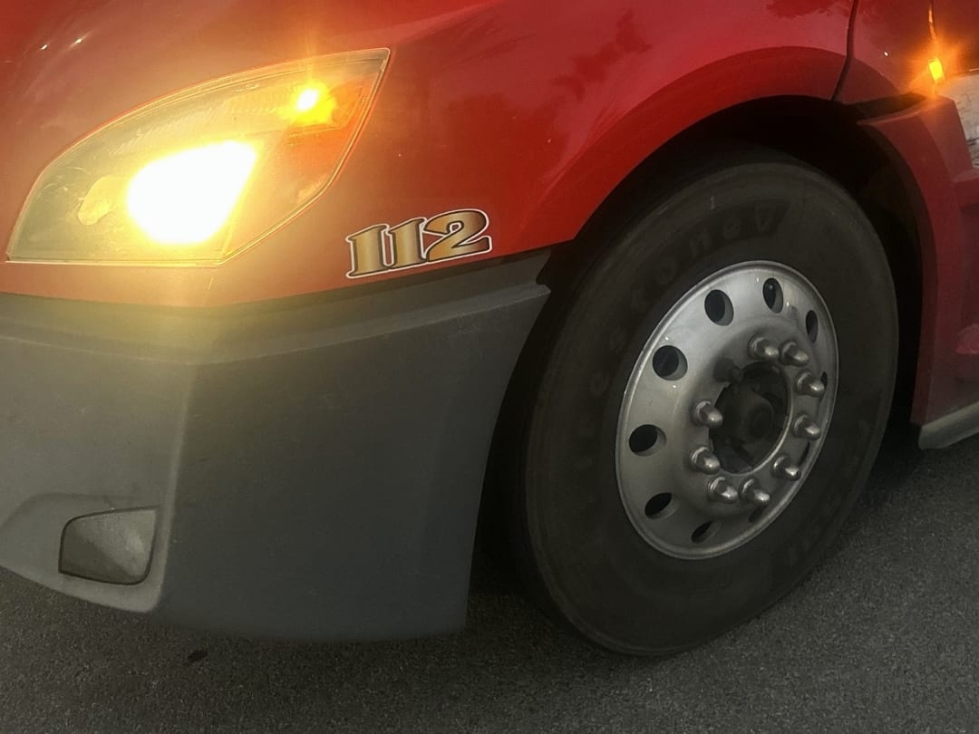 Unit 112, our red Cascadia, close up at dusk: headlight on, gold 112 numerals on the fender, front wheel.