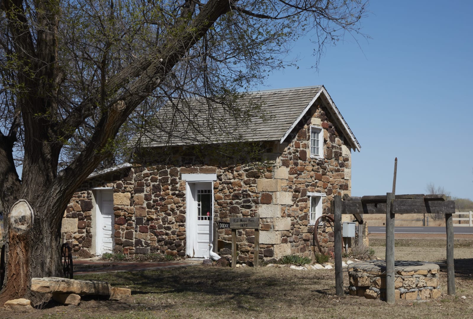 A small rough-stone house with white doors under a bare tree at the Barton County Historical Society village south of Great Bend.